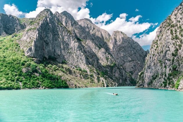 Ein kleines Boot fährt über türkisfarbenes Wasser in einer Schlucht, umgeben von steilen, felsigen Bergen mit grünen Hängen unter einem blauen Himmel mit weißen Wolken.
