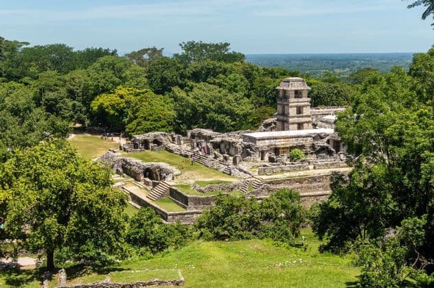 A wide-angle view of ancient stone ruins, including a prominent tower, surrounded by a dense green jungle under a clear sky.