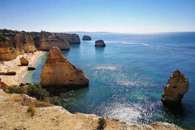 Un capo gruppo WeRoad con un cappello siede su una scogliera che si affaccia su una costa rocciosa con archi naturali sul mare e una piccola spiaggia.
