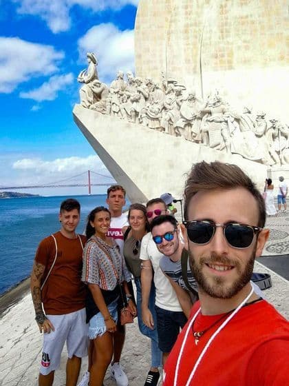 Un gruppo WeRoad si scatta un selfie davanti a un grande monumento in pietra in riva al mare, con un ponte sospeso in lontananza.
