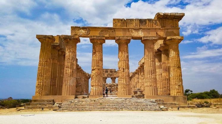 Les ruines d'un temple ancien avec des colonnes et des marches en pierre massives, avec deux personnes à l'intérieur sous un ciel bleu nuageux.