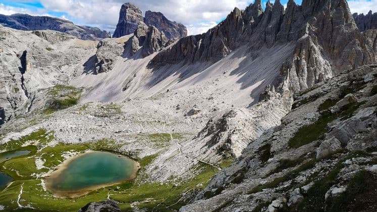 Veduta dall'alto di un lago alpino turchese e di un sentiero tortuoso in una vasta valle di montagna rocciosa, sotto un cielo parzialmente nuvoloso.
