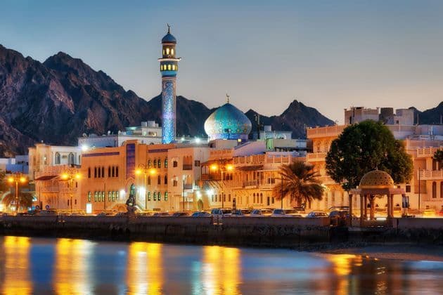 Una moschea illuminata e uno skyline cittadino su un lungomare al tramonto, con aspre montagne che si ergono dietro gli edifici.