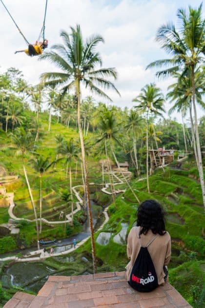 A person with a WeRoad backpack sits on a brick ledge, looking out at green rice terraces and palm trees as another person swings high above.