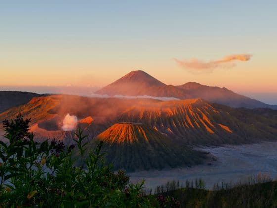 A landscape of volcanic craters illuminated by the golden light of sunrise, with one emitting a plume of smoke.
