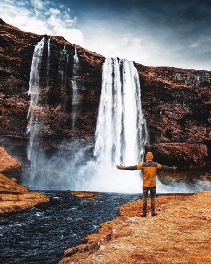 A person in a yellow jacket stands on a riverbank with arms outstretched, facing a large waterfall cascading down a rocky cliff.