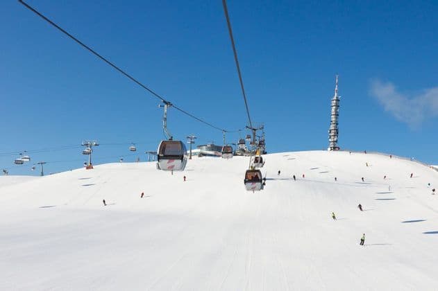 Seilbahnen fahren einen verschneiten Berg hinauf, während Skifahrer unter klarem blauem Himmel die Piste hinuntergleiten.