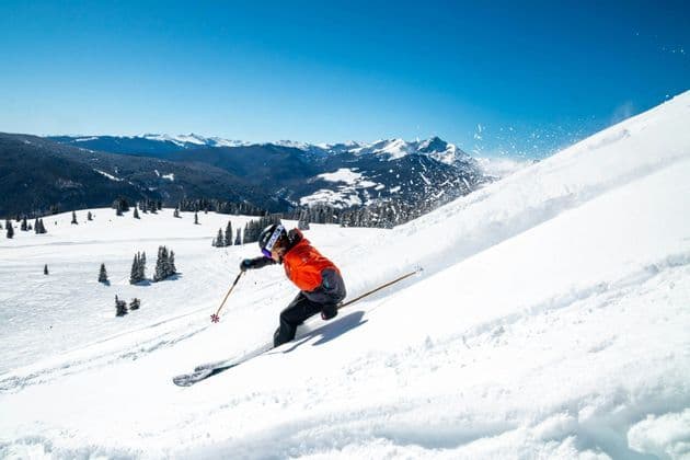 Ein Skifahrer in oranger Jacke carvt einen verschneiten Berghang hinunter und wirbelt Pulverschnee auf unter strahlend blauem Himmel.