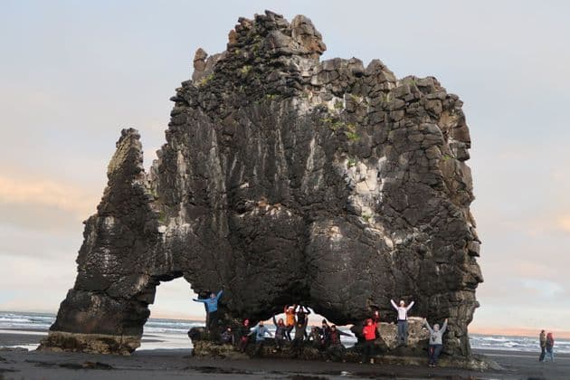 Un groupe WeRoad pose pour une photo sous une grande formation rocheuse arquée sur une plage de sable sombre.