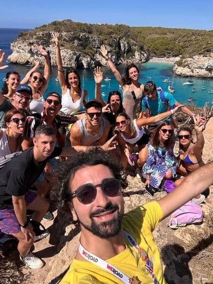 Eine WeRoad-Gruppe posiert für ein Selfie auf einer sonnigen Klippe, mit einer türkisfarbenen Bucht und Booten im Hintergrund.