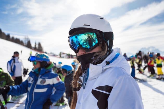 Ein Skifahrer mit einem weißen Helm und einer spiegelnden blauen Skibrille steht mit anderen Skifahrern auf einer schneebedeckten Skipiste.