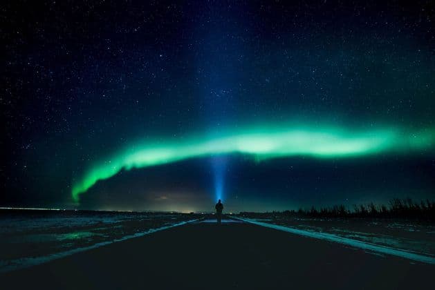 A person stands on a dark road, shining a flashlight up at the green aurora borealis under a starry night sky.