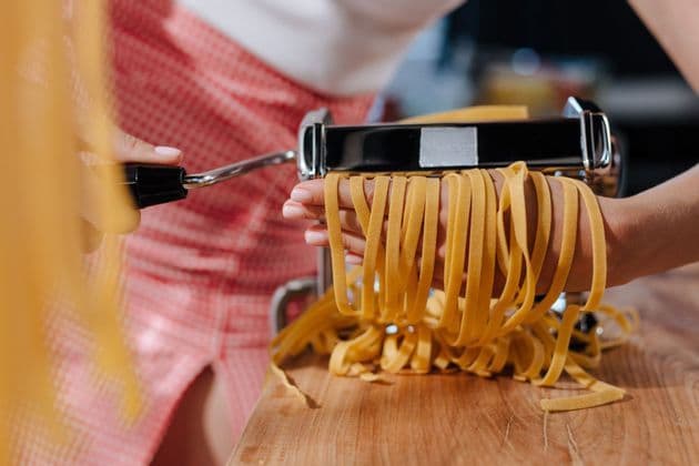 A person's hands catch fresh tagliatelle as it comes out of a manual pasta maker on a wooden surface.