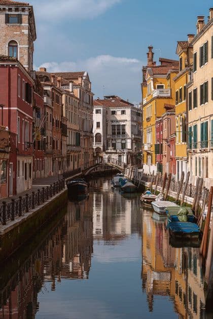 A view down a narrow canal lined with colorful buildings, whose reflections are visible in the water alongside moored boats.