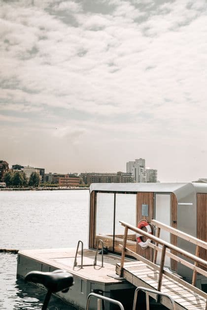 A floating sauna cabin with a wooden deck and walkway sits on an urban waterfront under a cloudy sky.