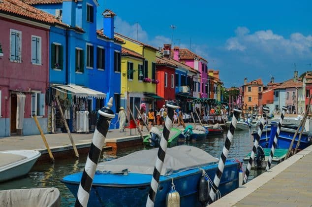 Boats are moored in a canal next to a walkway lined with brightly colored buildings under a clear blue sky.