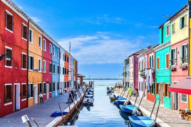 Brightly colored houses line a canal filled with small moored boats, leading out to the sea under a blue sky.