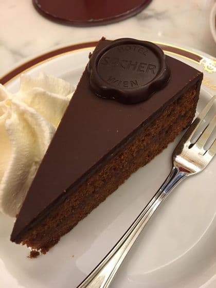 Una porción de tarta Sacher con su sello de chocolate, servida en un plato blanco con nata montada al lado y un tenedor.