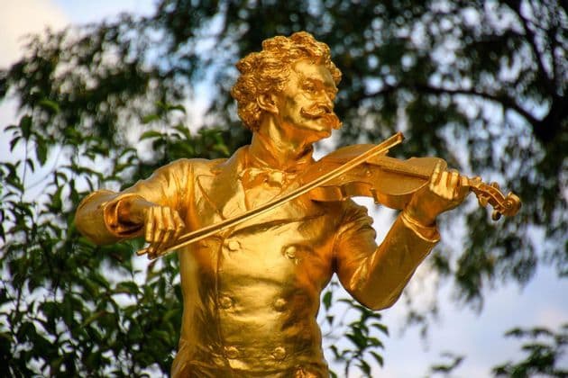 Una estatua dorada de un hombre con bigote tocando un violín, con árboles verdes difuminados al fondo.