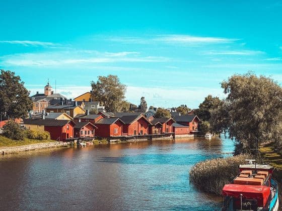 Une rangée de maisons en bois rouges borde la rive d'une rivière calme, avec des arbres et une ville en arrière-plan sous un ciel bleu.