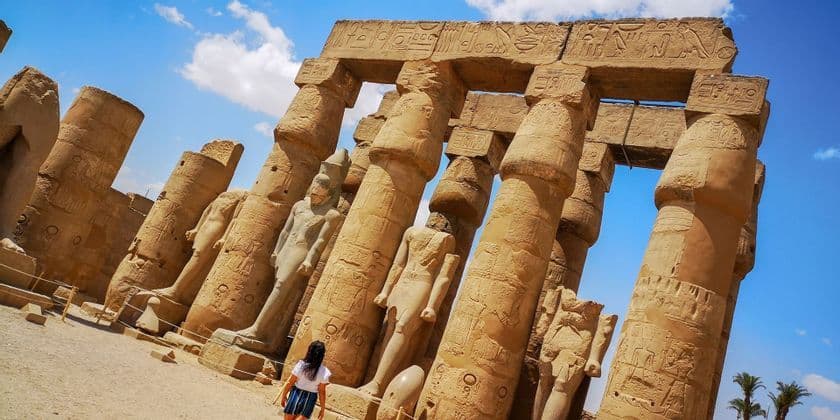 Eine Frau steht da und blickt unter einem leuchtend blauen Himmel zu überragenden antiken Steinsäulen und Statuen auf.