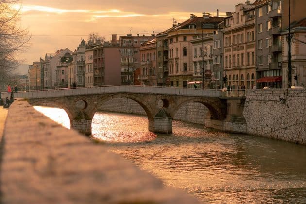Un puente de piedra en arco cruza un río que fluye por una ciudad, con la luz dorada del atardecer reflejándose en el agua.