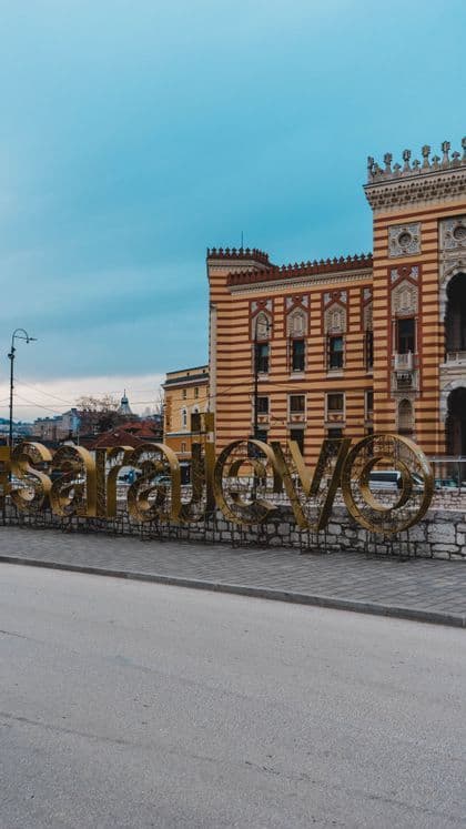 Una grande insegna dorata 'Sarajevo' si erge su un muro di pietra davanti a un edificio a strisce ornato sotto un cielo nuvoloso.