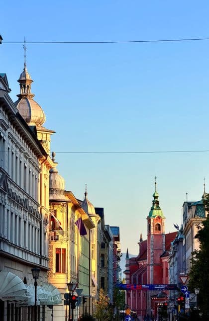 Ornate city buildings and a distant church steeple are lit by warm sunlight against a clear blue sky.