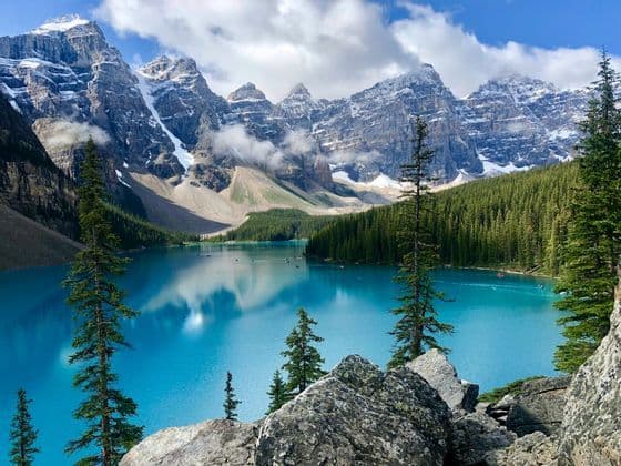 Una vista su un lago alpino turchese verso montagne frastagliate e innevate, con una foresta sempreverde lungo la riva.
