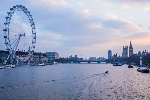 Una grande ruota panoramica si erge sulla riva di un fiume con barche che navigano accanto agli edifici della città al crepuscolo