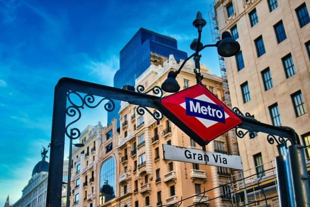 Un'insegna d'ingresso di una stazione della metropolitana d'epoca con la scritta Gran Via sullo sfondo di classici edifici cittadini e un cielo blu luminoso.