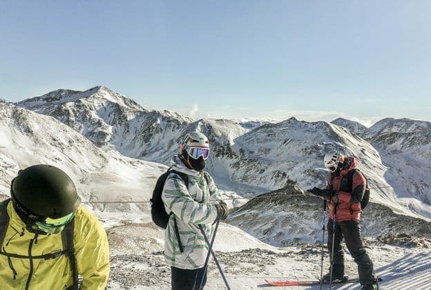 Un voyage de groupe WeRoad de skieurs en tenue debout au sommet d'une montagne enneigée avec des pics en arrière-plan