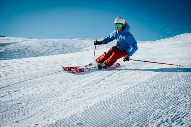 Un sciatore con giacca blu e pantaloni rossi che scende con curve condotte lungo un pendio innevato e battuto in una giornata di sole.