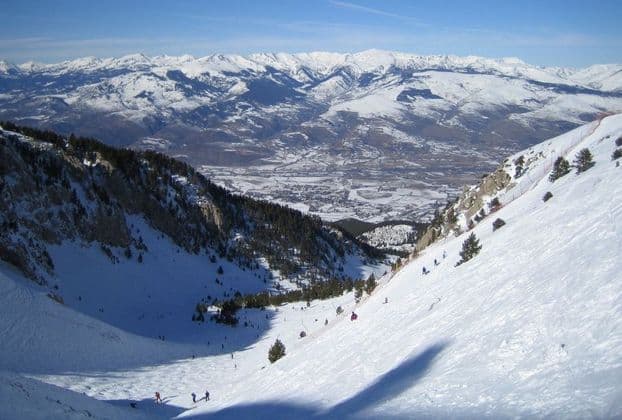 Esquiadores descienden por una empinada pendiente nevada con vistas a un valle y picos lejanos bajo un cielo azul.