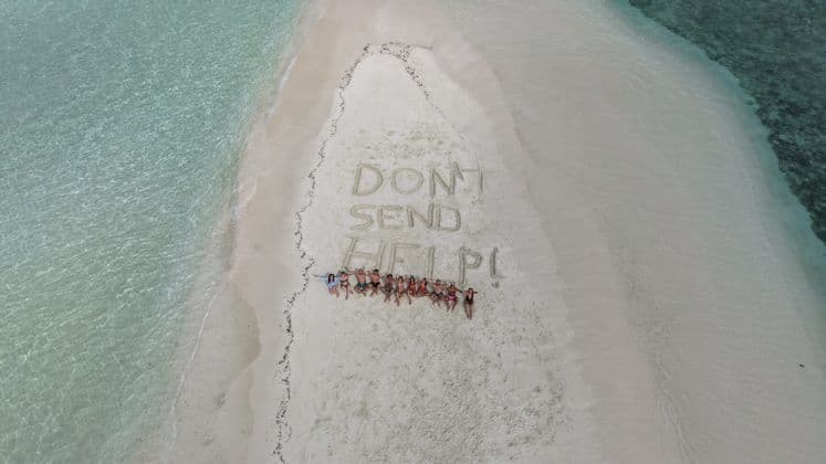 Un viaggio di gruppo WeRoad sdraiati su un banco di sabbia accanto alla scritta 'NON MANDATE AIUTO!' tracciata nella sabbia e circondata da acqua cristallina.
