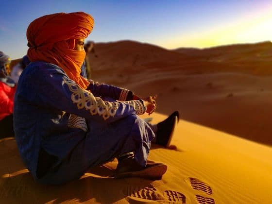 Una persona en un viaje grupal de WeRoad, con un turbante naranja, se sienta en una duna de arena observando el atardecer en el desierto.