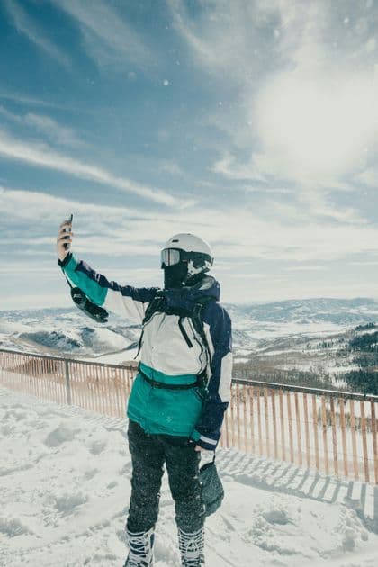 Un snowboarder con attrezzatura invernale completa si fa un selfie su una cima montuosa innevata in una giornata di sole.