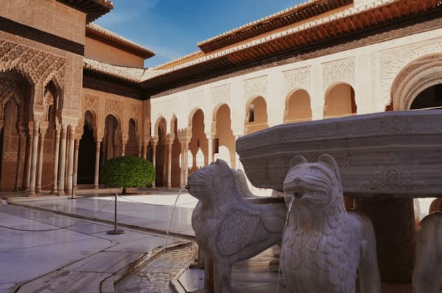 Una fuente de piedra sostenida por estatuas de leones esculpidas que arrojan agua en un patio ornamentado con arcos detallados.