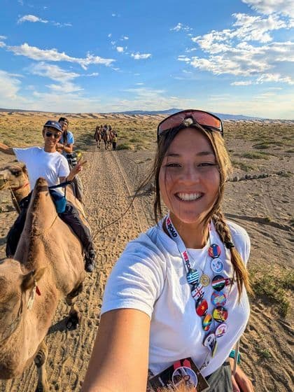 Un groupe WeRoad souriant qui fait une balade à dos de chameau sur un sentier de sable à travers un paysage désertique ensoleillé