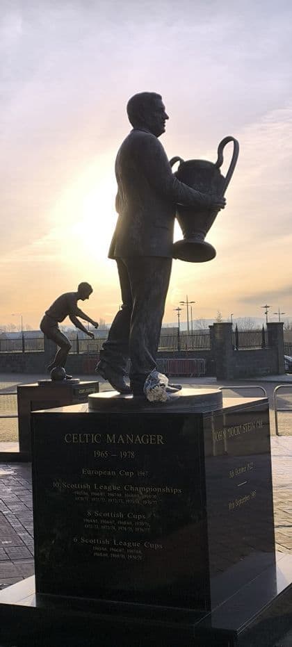 Statua in bronzo di un allenatore di calcio che tiene un trofeo su un piedistallo contro un cielo al tramonto