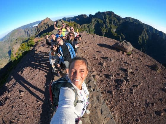 Un voyage en groupe WeRoad sourit pour un selfie sur une crête rocheuse de montagne avec des sommets en arrière-plan.