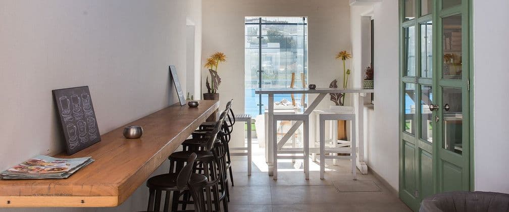 Luminoso interno di un bar con un lungo bancone in legno, sgabelli da bar e grandi finestre che si aprono su un'area esterna.