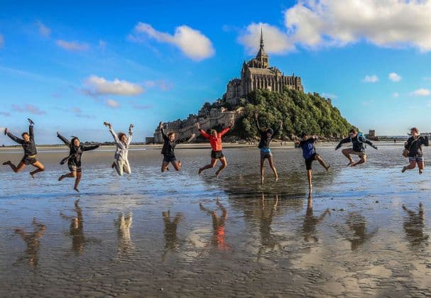 Eine WeRoad-Gruppe springt gemeinsam an einem nassen Sandstrand, ihre Spiegelungen im Wasser sichtbar und eine Burg auf einem Hügel im Hintergrund.