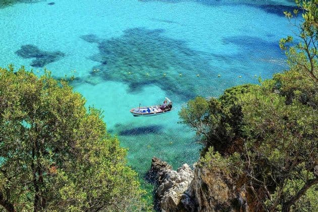 A small boat with passengers floats on transparent turquoise sea, viewed from a cliffside covered with green trees.