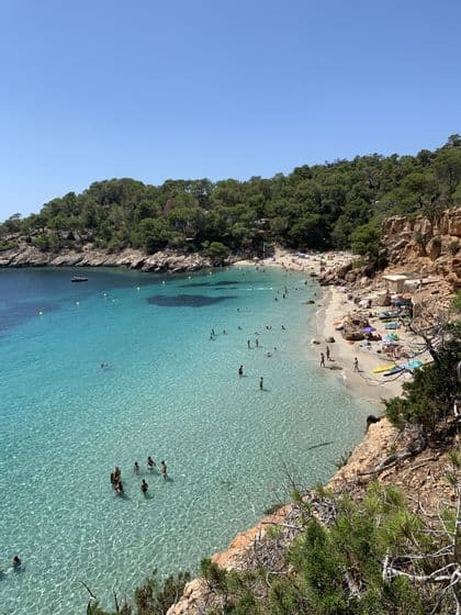 Una vista dall'alto di una caletta affollata con persone che nuotano in acqua limpida e turchese e prendono il sole sulla sabbia.