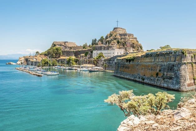 A stone fortress on a hill overlooks a marina with sailboats docked in a bay with turquoise water on a sunny day.