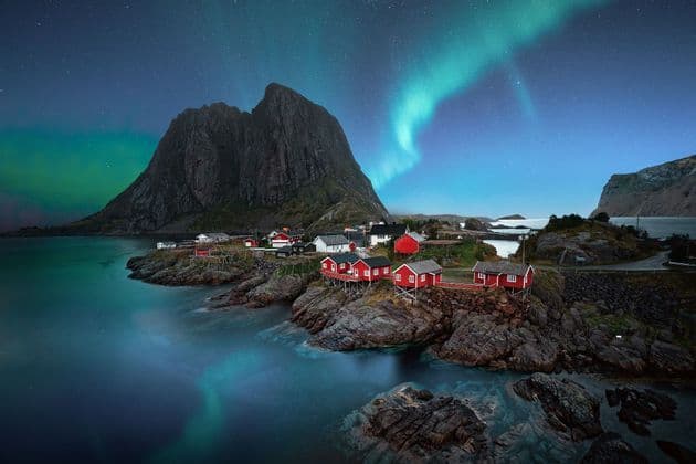 Red stilt houses on a rocky coast below a large mountain, with the green Northern Lights visible in the starry night sky.
