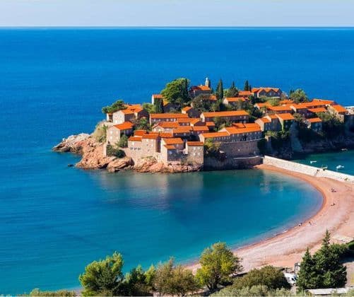 Un piccolo borgo insulare fortificato con tetti in tegole arancioni è circondato dal mare azzurro e collegato a una spiaggia di sabbia curva da una strada rialzata.
