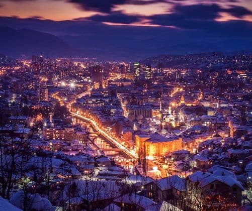 Una vista elevata di una città innevata illuminata dalle luci al crepuscolo, con montagne in silhouette sullo sfondo.