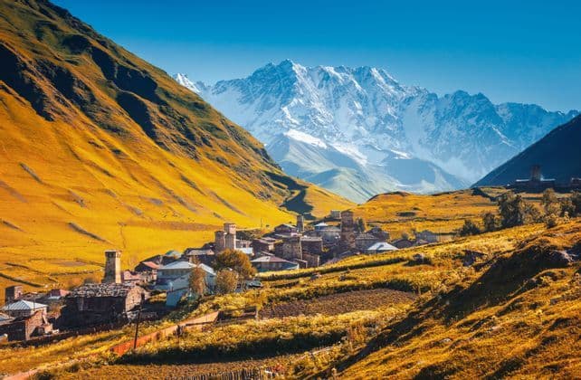 Pueblo de piedra con torres medievales enclavado en un valle bajo picos de montañas nevados bajo un cielo azul despejado